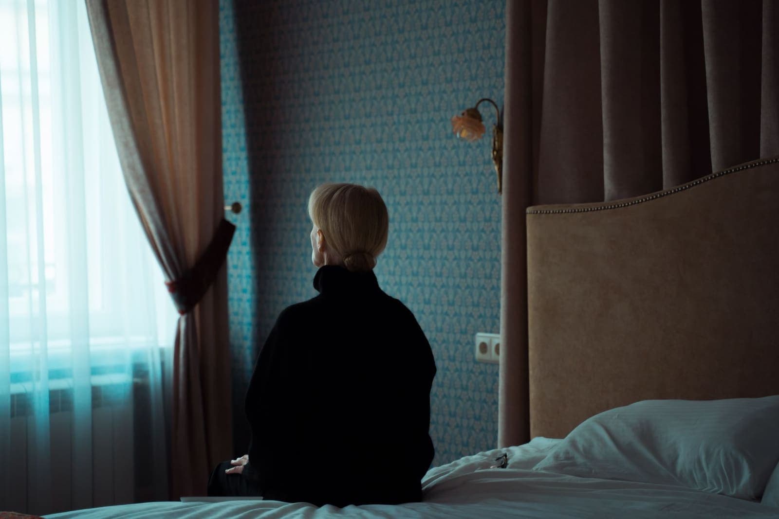 Femme assise au bord de son lit face à la fenêtre dans la pénombre, réveil nocturne